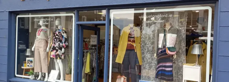 A clothing shop window display featuring mannequins dressed in colourful outfits, including skirts, trousers, tops, and jackets. The shop’s name and website appear on the window above the mannequins.