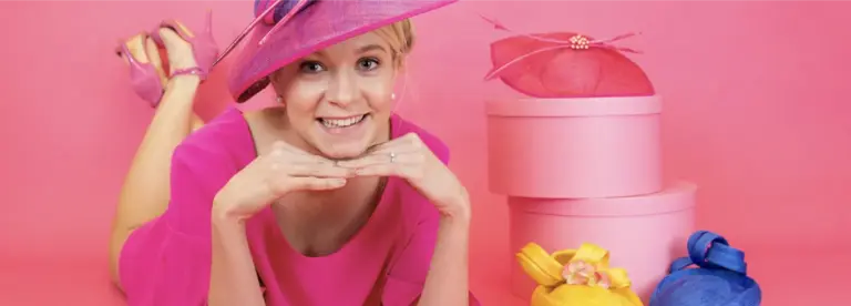 A person in a bright pink dress and wide-brimmed hat smiles whilst lying on the floor with hands under their chin. Pink hat boxes and colourful hats in yellow and blue are arranged nearby against a pink background.