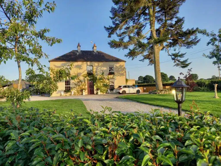 A large yellow two-storey house with a dark roof stands in a manicured garden with green grass, tall trees, a gravel drive, and a white car parked beside the house on a sunny day. A lamp and bushes are in the foreground.