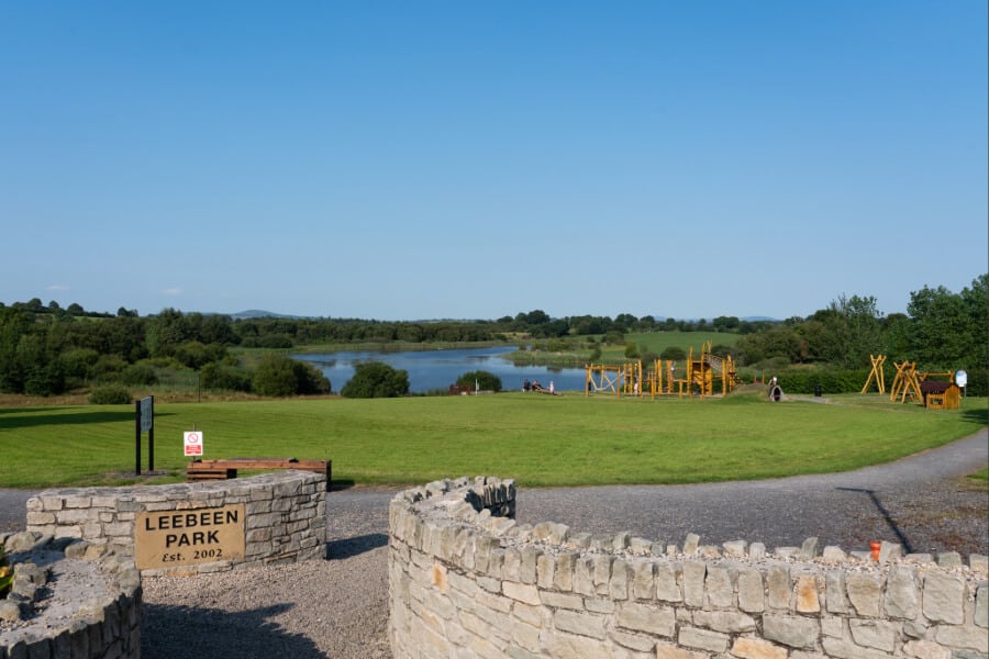 Entrance to Leebeen Park with a stone wall and sign, a large grassy field, playground equipment, people walking, and a pond surrounded by trees under a clear blue sky.