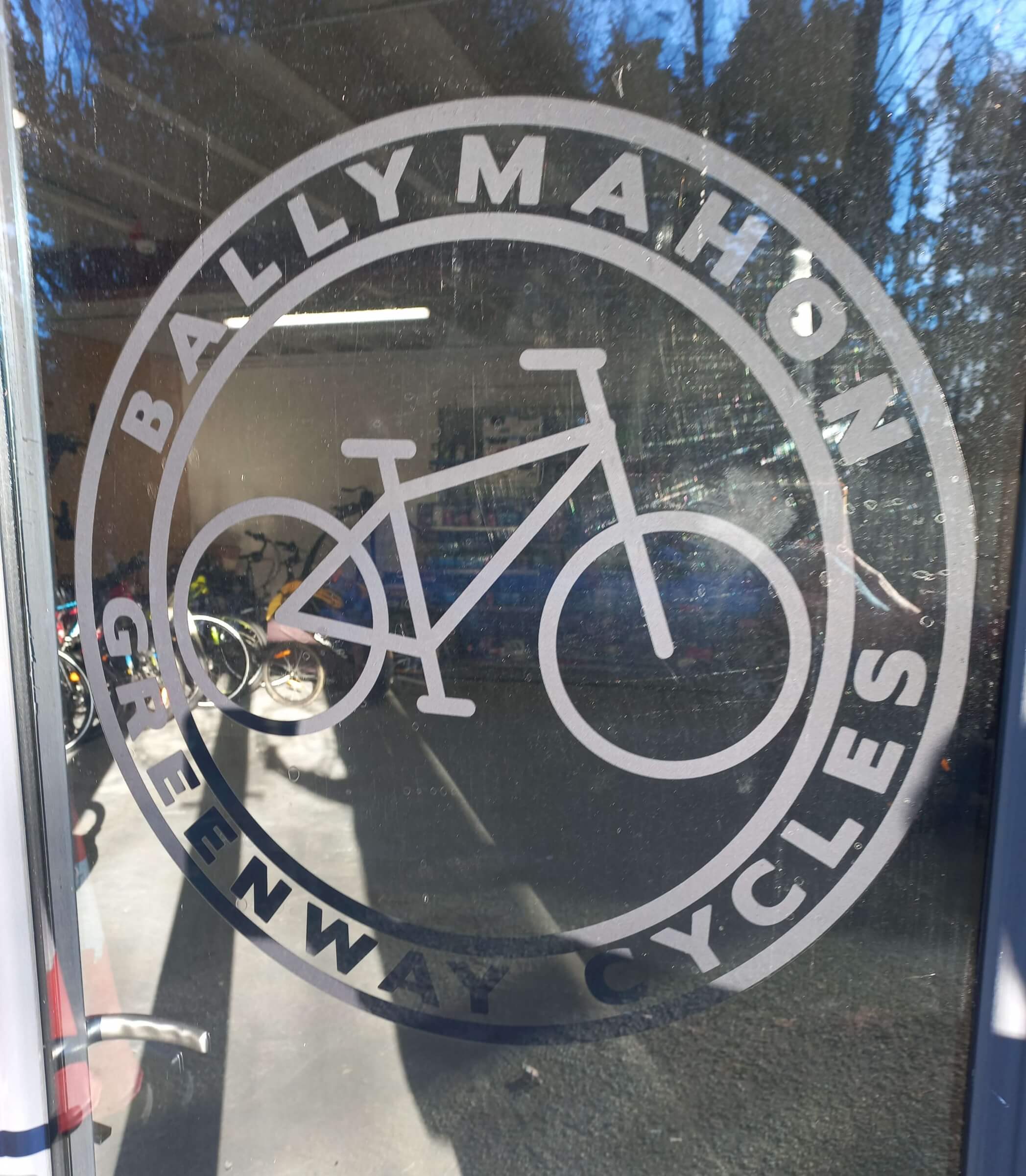 A circular logo on glass reads "Ballymahon Greenway Cycles" and features a simple bicycle graphic in the centre. Reflections and bicycles are visible through the glass.