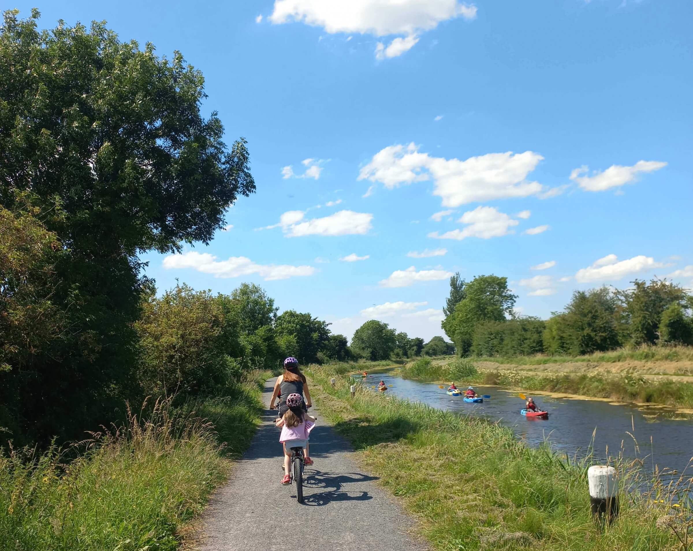 A woman and a child ride bicycles on a path beside a river, while several people kayak on the water under a blue sky with fluffy clouds. Trees and grass line both sides of the path and river.