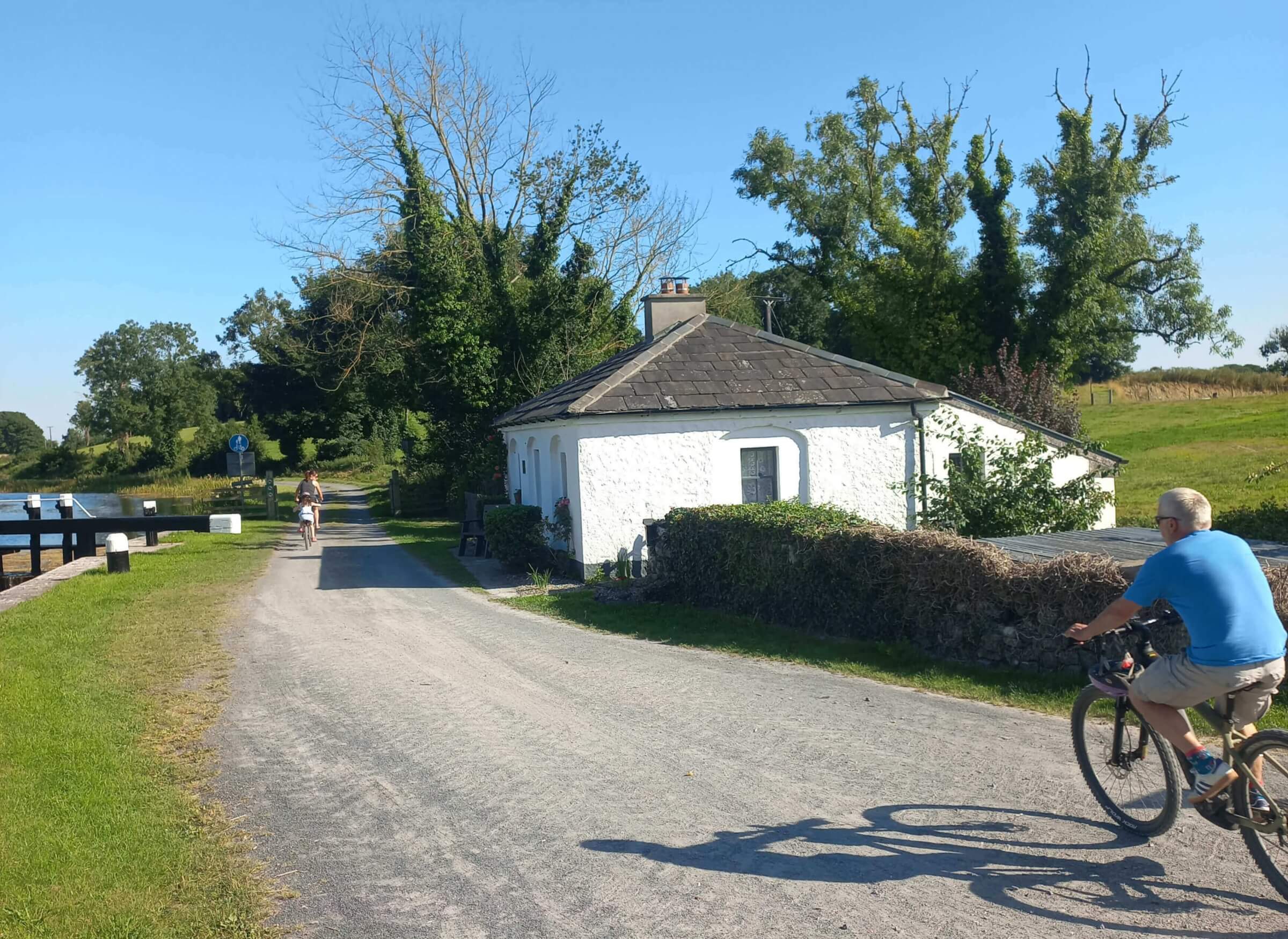 Two people ride bicycles along a gravel path beside a white cottage and a canal on a sunny day, with trees and grass bordering the scene.
