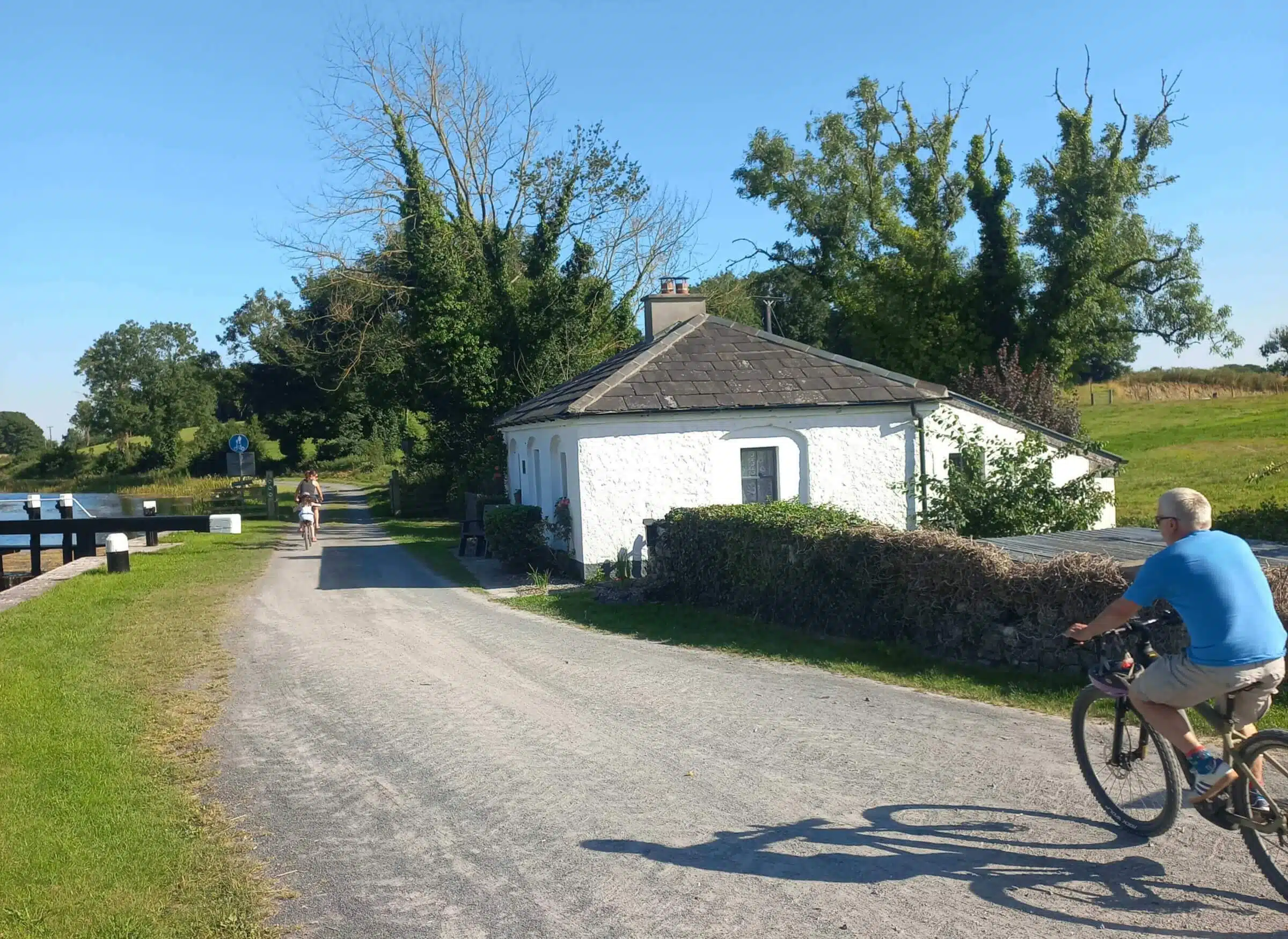 Two people ride bicycles along a gravel path beside a white cottage and a canal on a sunny day, with trees and grass bordering the scene.
