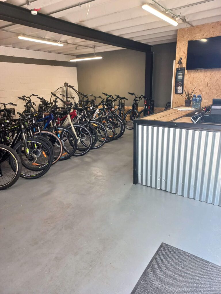 A bike shop interior with several rows of bicycles lined up against a wall, a large fan, and a counter with a TV, water bottles, and a first aid kit on the wall. The floor and walls are clean and industrial in style.