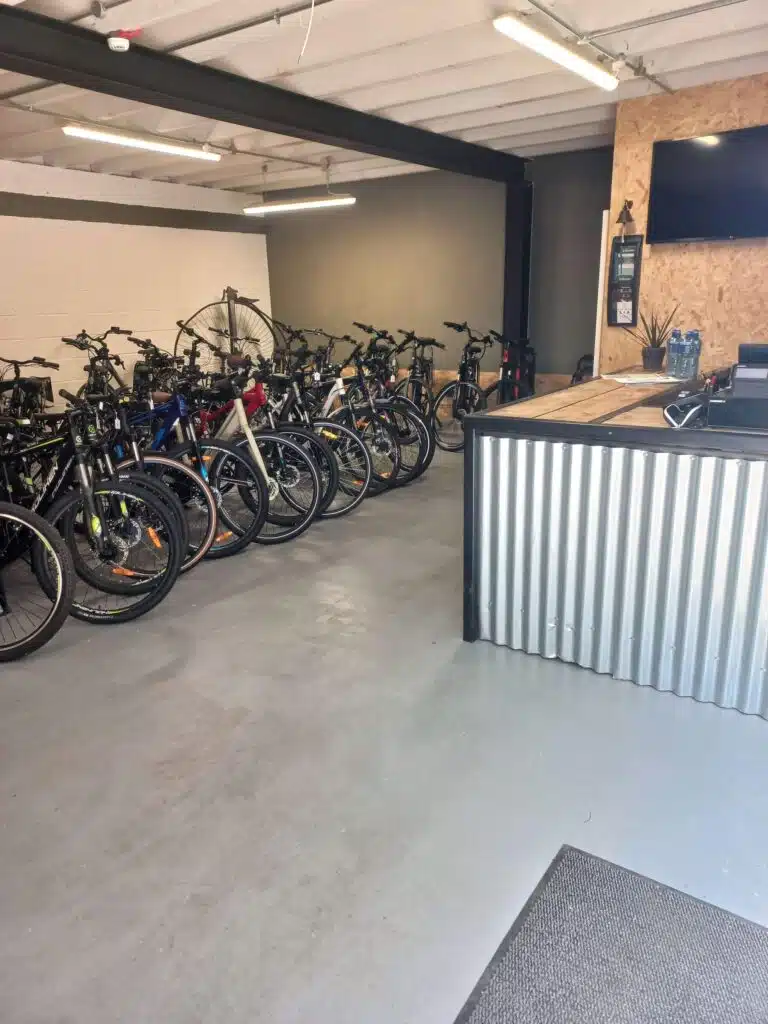 A bike shop interior with several rows of bicycles lined up against a wall, a large fan, and a counter with a TV, water bottles, and a first aid kit on the wall. The floor and walls are clean and industrial in style.