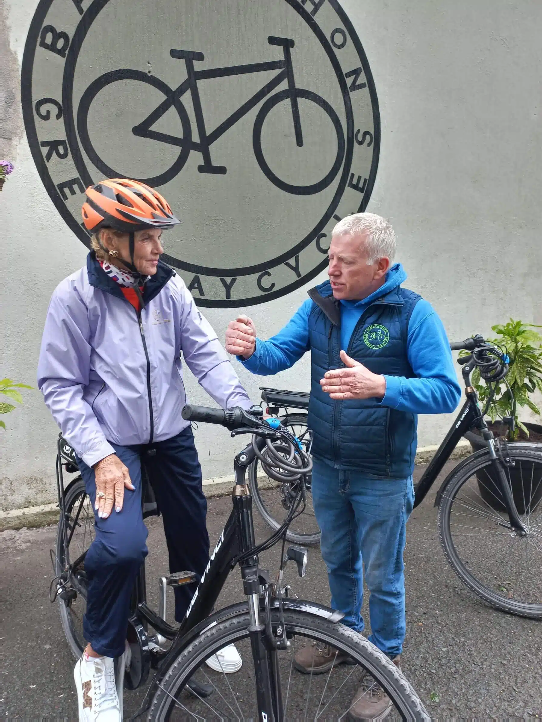An older person wearing a helmet sits on a bicycle, whilst another person stands beside them gesturing and talking. They are outside in front of a wall with a large bicycle graphic and the words "Greenway Cycles.