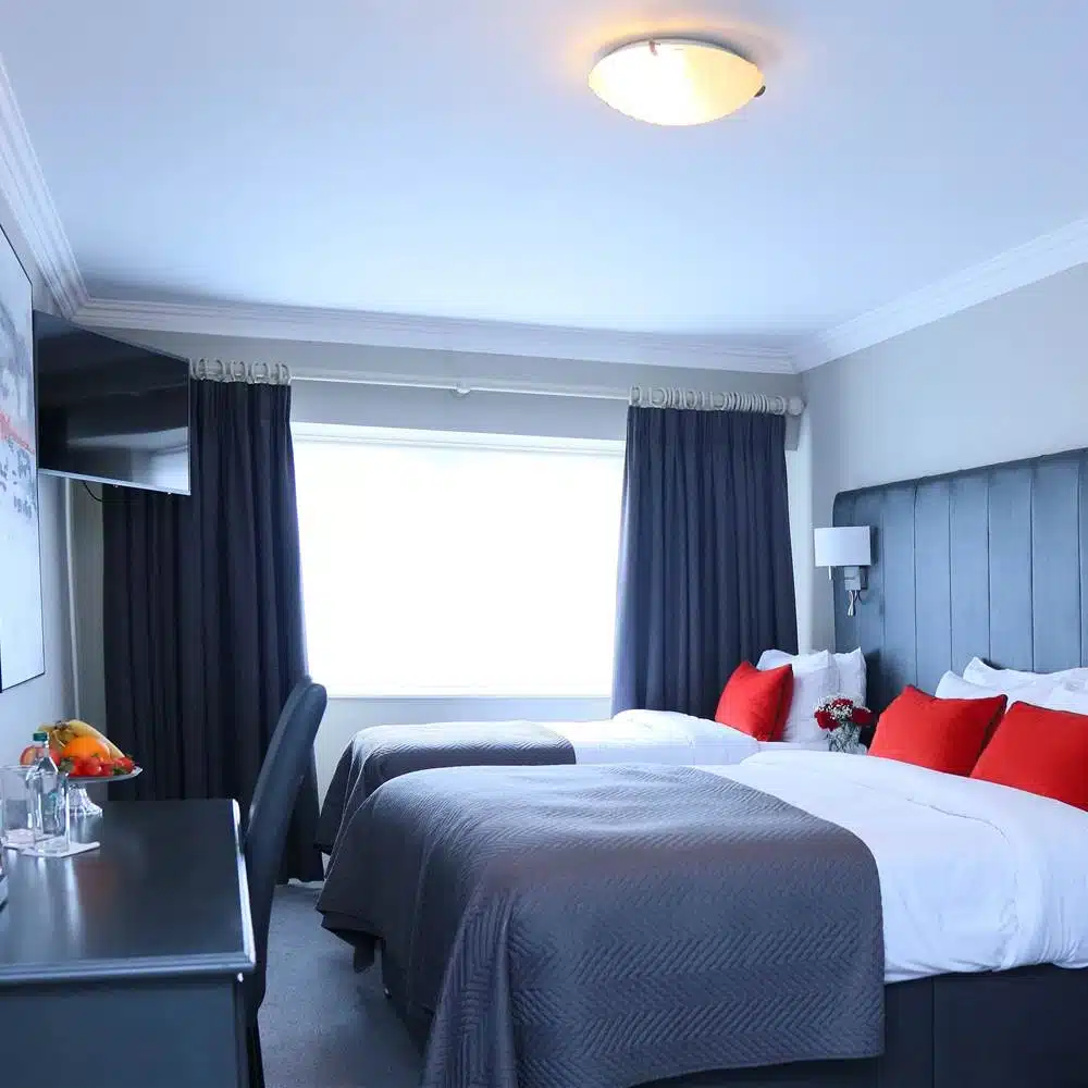 A modern hotel room with two beds, grey headboards, grey bedding, and red accent cushions. There is a desk with a chair, fruit bowl, water bottles, and a flat-screen TV on the wall. Large window with dark curtains.