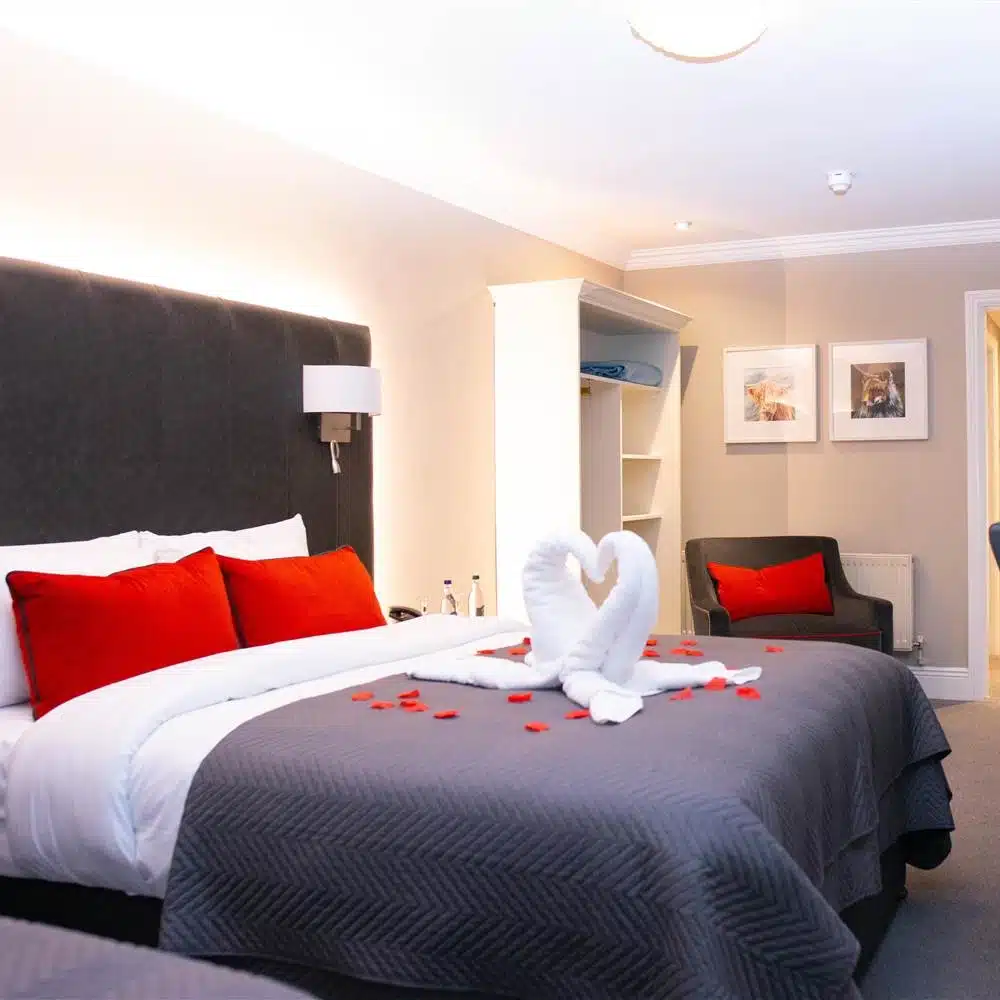 A hotel room with a large bed, grey headboard, grey bedding, and red pillows. Towel swans form a heart shape on the bed, surrounded by rose petals. An armchair, artwork, and open shelving are visible in the background.