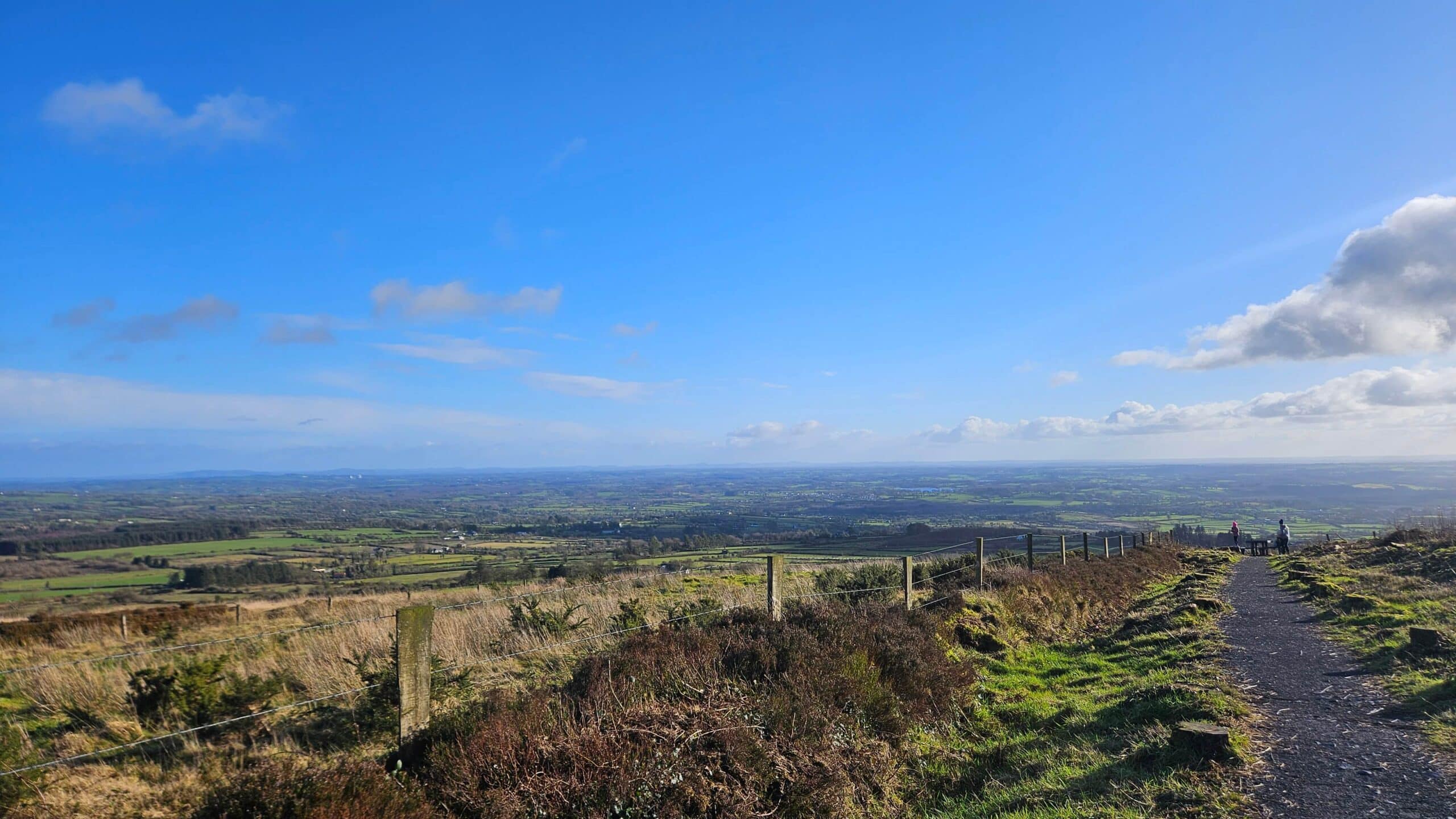 A scenic view from a hilltop shows a dirt track and fence running along grassy terrain, overlooking wide green fields under a bright blue sky with scattered clouds.