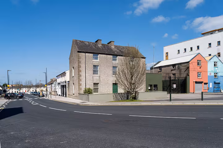 A quiet street corner features an old, grey, two-storey house with a green door, surrounded by a low wall. Nearby are colourful buildings and a clear blue sky with scattered clouds.