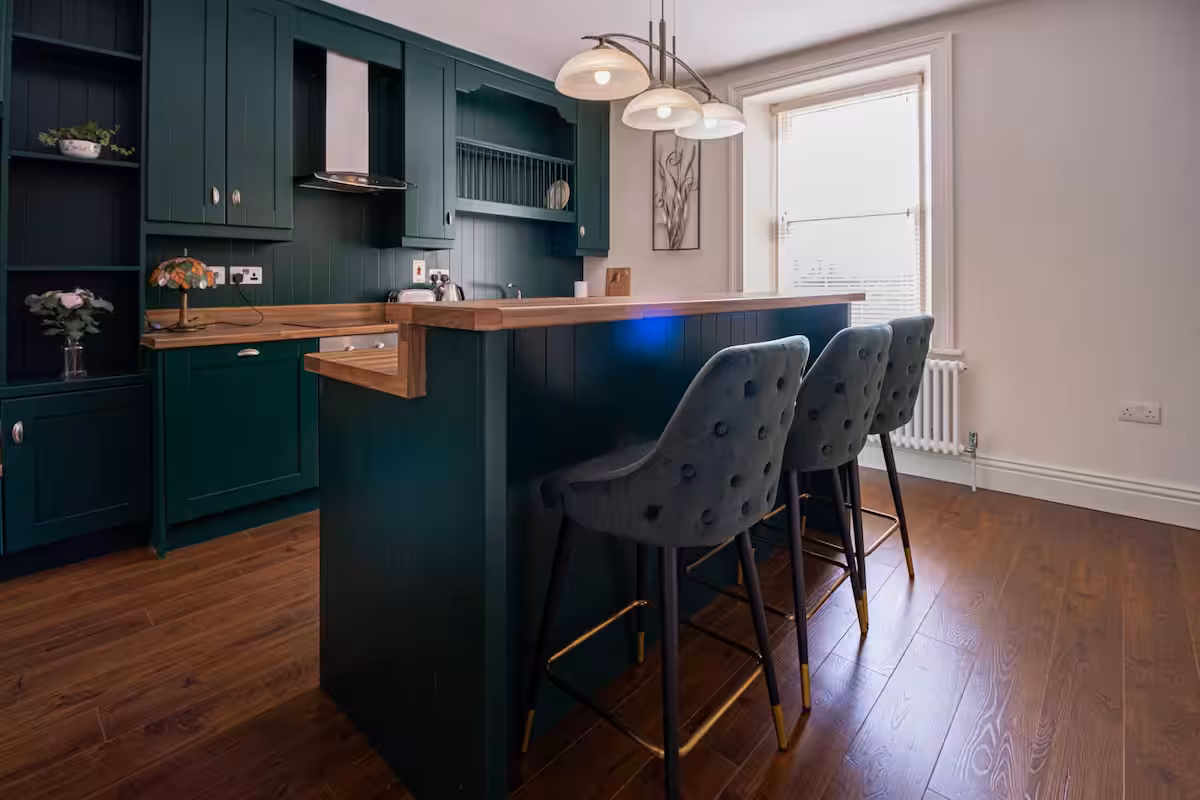 Modern kitchen with dark green cupboards, a wooden island, three grey upholstered bar stools, wooden floors, pendant lighting, and a large window letting in natural light.