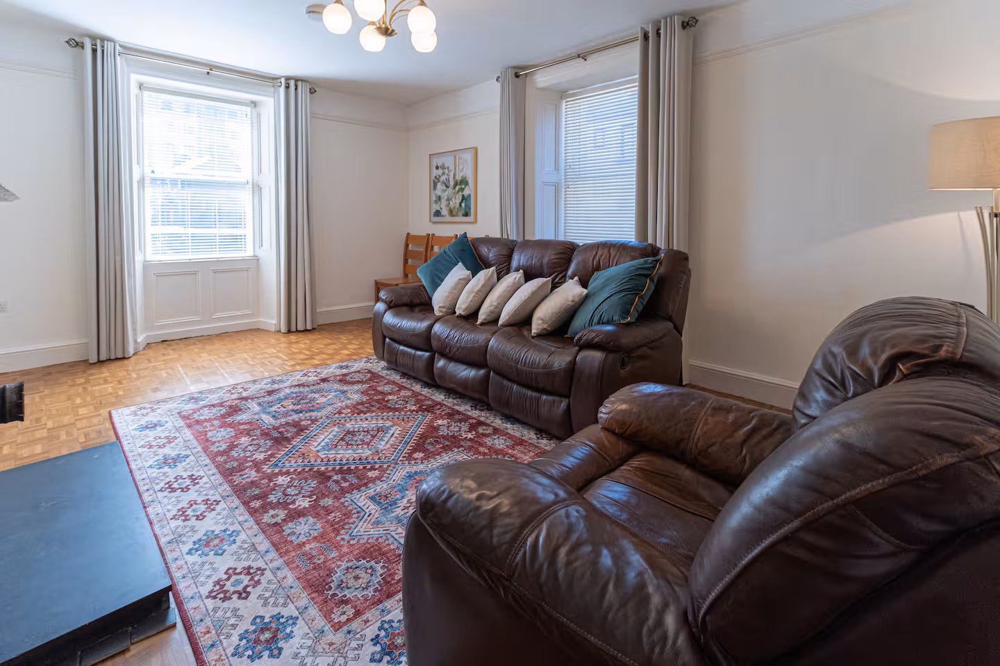 A cosy living room with a brown leather sofa and armchair, colourful patterned rug, two windows with curtains, a standing lamp, a framed wall picture, and a wooden chair in the corner.