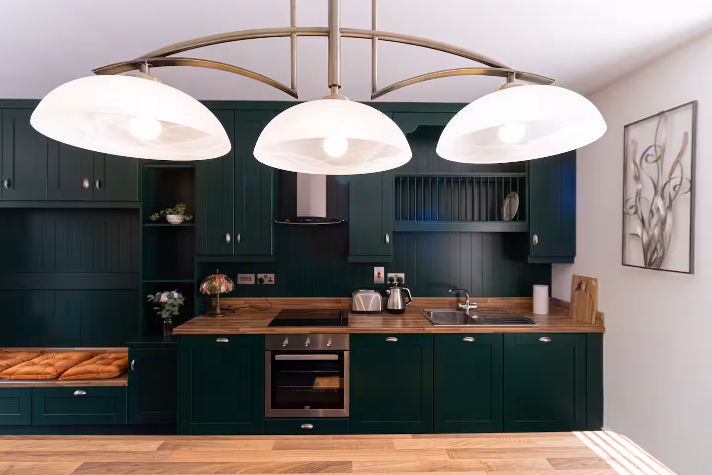 Modern kitchen with dark green cupboards, wooden worktops, and a built-in oven. Three large white pendant lights hang above. A sink, kettle, toaster, wall art, and a cushioned bench are also visible.