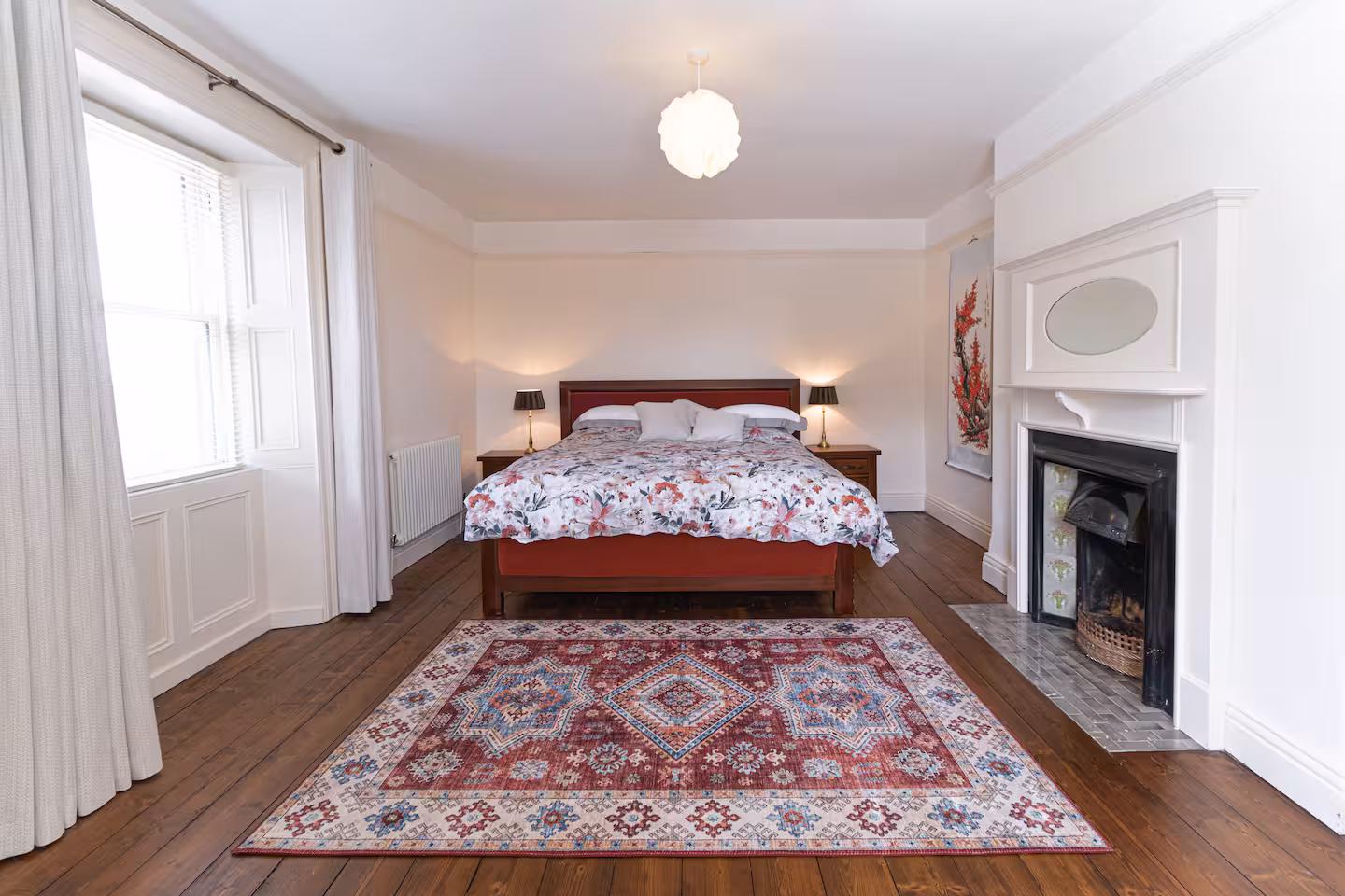 A bright bedroom with a double bed, floral bedding, two bedside tables with lamps, a fireplace, wooden floor, a patterned rug, and a large window with white curtains.