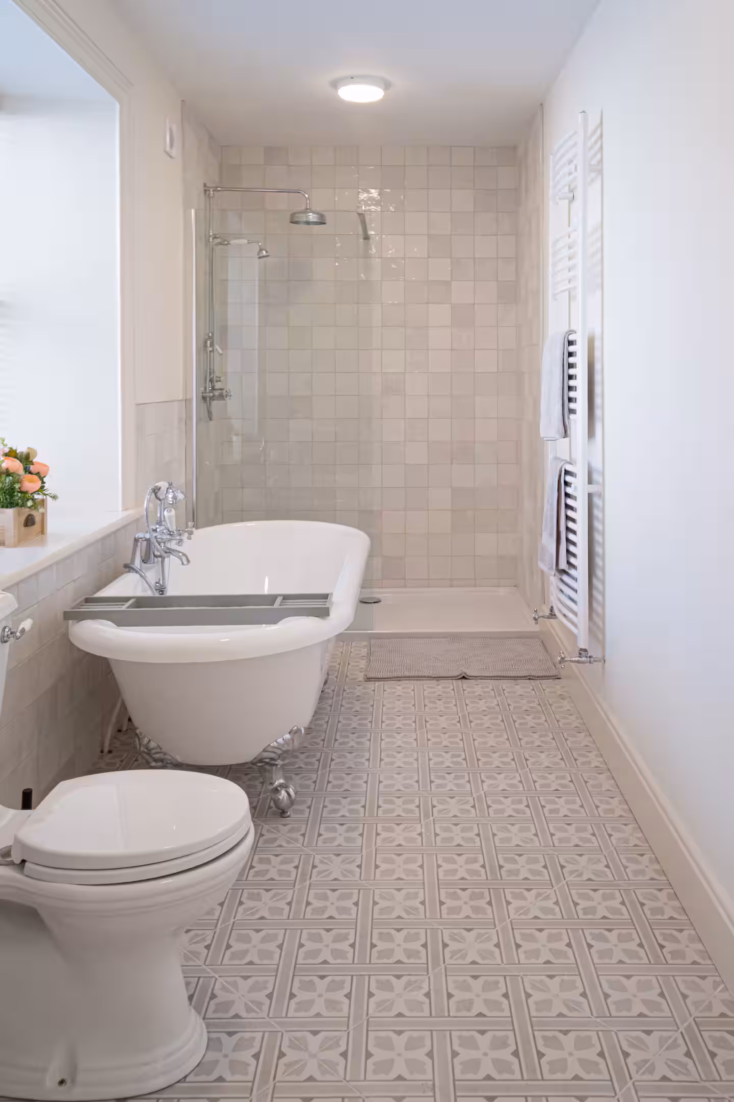 A bright, modern bathroom with patterned tiled flooring, a white clawfoot bath, a toilet, a walk-in shower with glass doors, and white walls. A small potted plant sits by the bath.