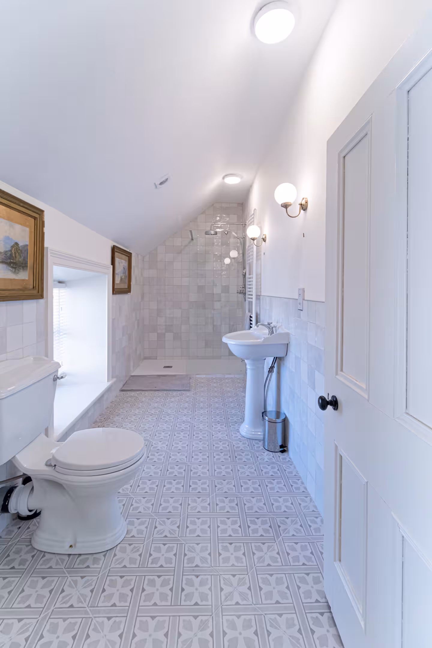 Bright bathroom with white patterned floor tiles, sloped ceiling, toilet, pedestal basin, shower area at the far end, wall-mounted lights, a window, and framed pictures on the walls.
