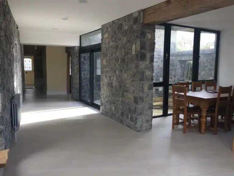 A modern dining area with stone walls, large windows, a wooden dining table with chairs, and tiled floors. Sunlight streams in through the windows, creating a bright and airy space.