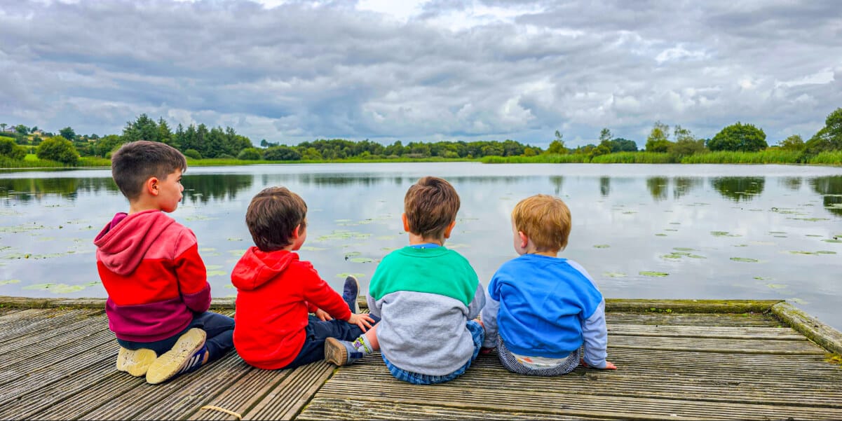 Four young children sit on a wooden jetty, facing a calm lake surrounded by greenery under a cloudy sky. The children wear colourful jackets and appear to be enjoying the peaceful nature scene.
