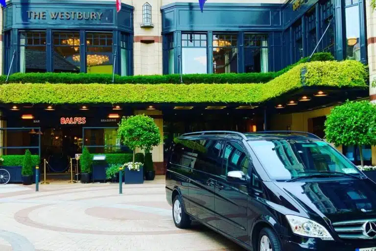 A black van is parked in front of The Westbury hotel entrance, with green hedges, potted plants, and large windows above the doorway. The Balfe’s restaurant sign is visible near the entrance.