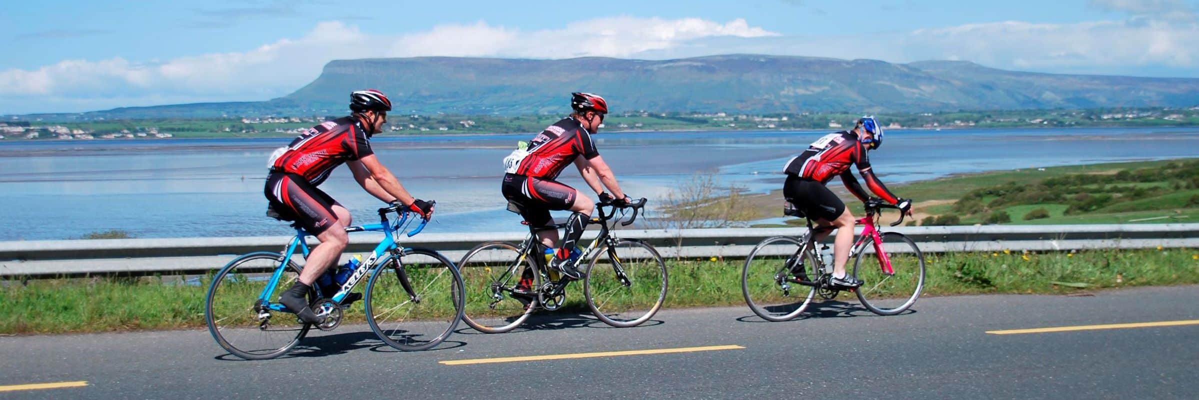Three cyclists in matching red and black gear ride on a tarmacked road beside a scenic body of water, with green fields and distant mountains under a partly cloudy sky in the background.