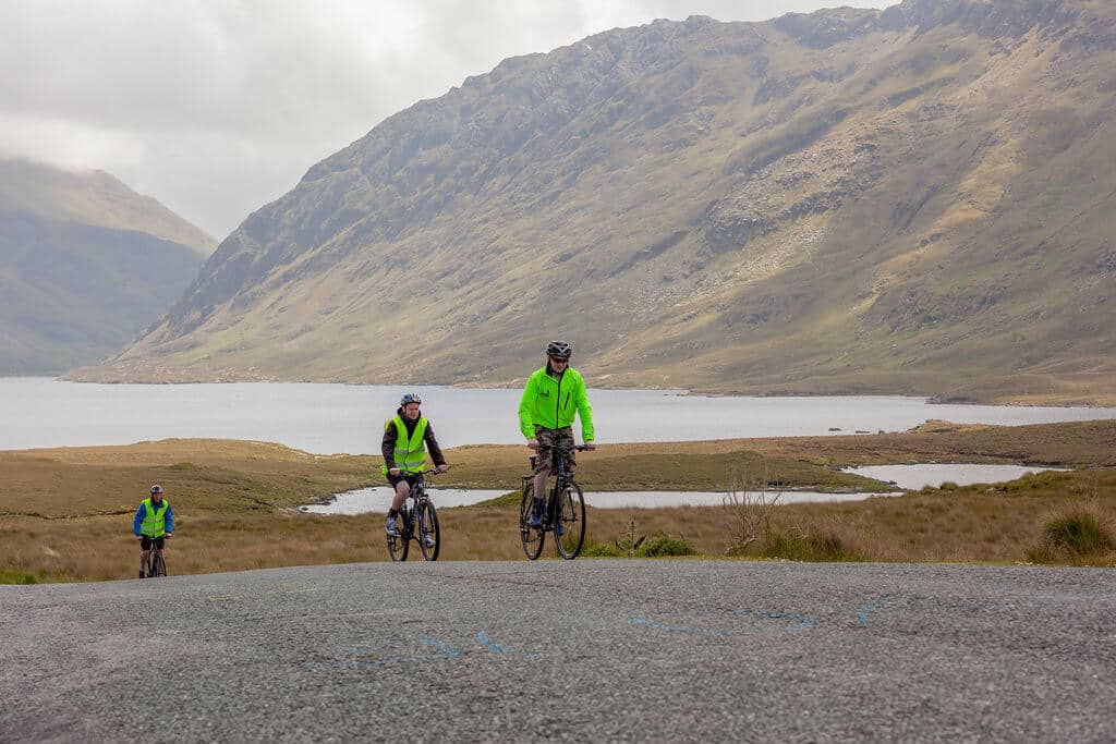 Three cyclists wearing bright green jackets ride on a tarmacked road with a scenic backdrop of mountains, a lake, and grassy terrain under a cloudy sky.