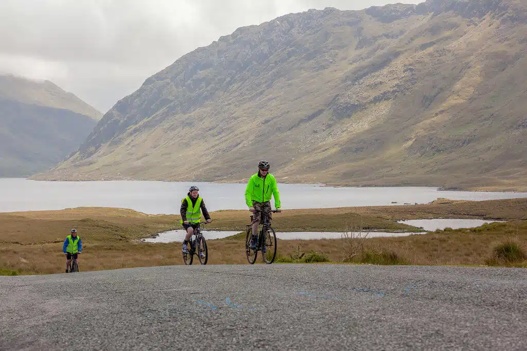 Three cyclists wearing bright green jackets ride on a tarmacked road with a scenic backdrop of mountains, a lake, and grassy terrain under a cloudy sky.