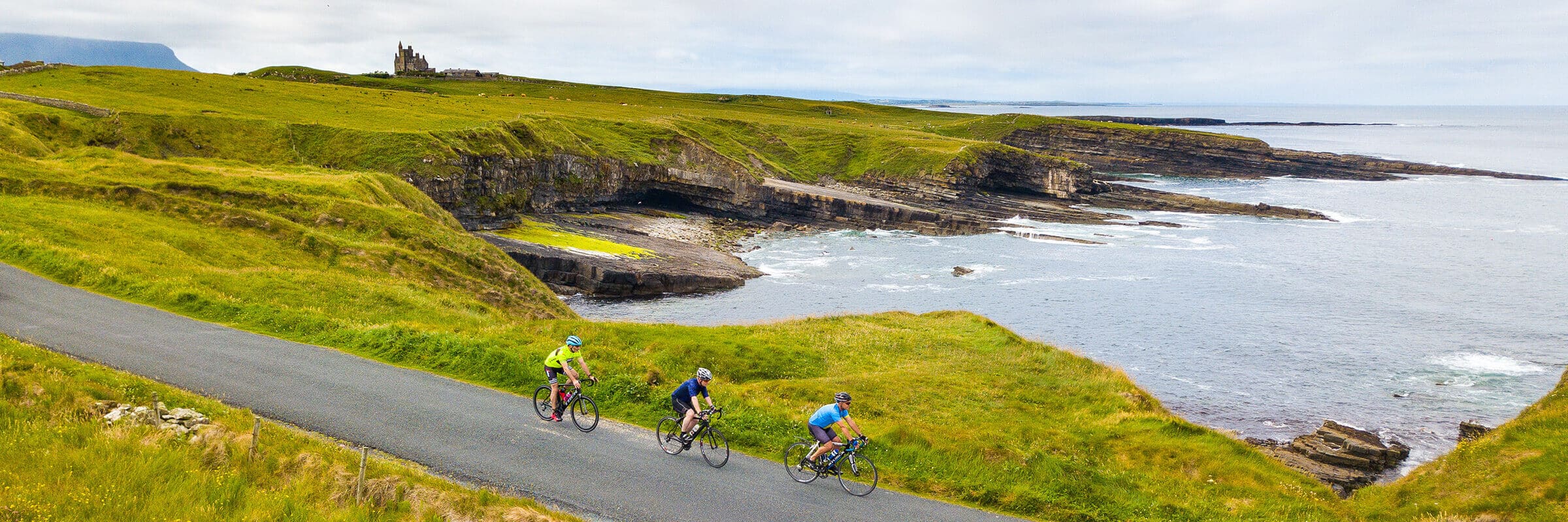 Three cyclists ride along a coastal road with green cliffs, rocky shoreline, and the sea on one side. An old castle stands atop the distant hill under a cloudy sky.