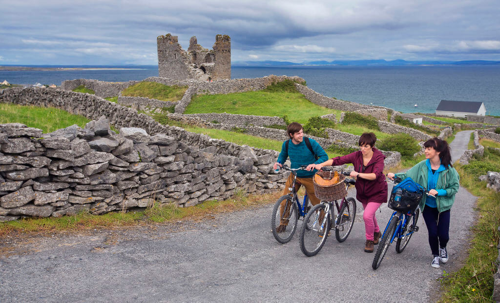 Three people walk their bicycles up a tarmacked road, bordered by stone walls, with a ruined stone castle and the sea in the background under a cloudy sky.