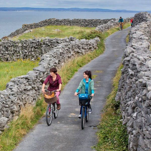 Two people ride bicycles along a narrow tarmac path bordered by stone walls, with grassy fields, the sea, and distant hills in the background. Other cyclists are visible further along the path.