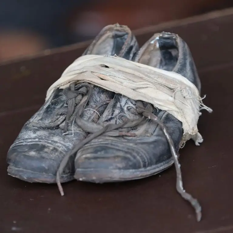 A pair of old, worn-out black shoes with frayed laces are wrapped together with a tattered white cloth, sitting on a dark surface.