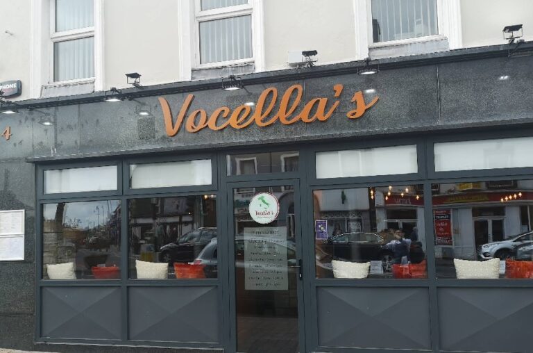 Street view of "Vocella's" restaurant with black and grey exterior, large front windows, and orange sign above the entrance. Reflections and parked cars can be seen through the windows.
