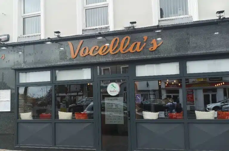 Street view of "Vocella's" restaurant with black and grey exterior, large front windows, and orange sign above the entrance. Reflections and parked cars can be seen through the windows.