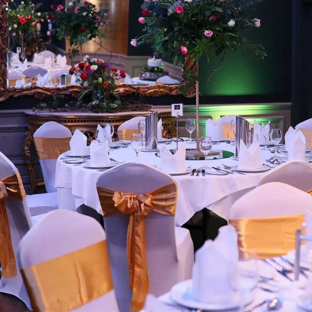A round dining table set for a formal event with white tablecloth, neatly folded serviettes, glassware, and cutlery. Chairs have white covers with gold sashes. Floral and greenery centrepieces are visible in the elegant, decorated room.