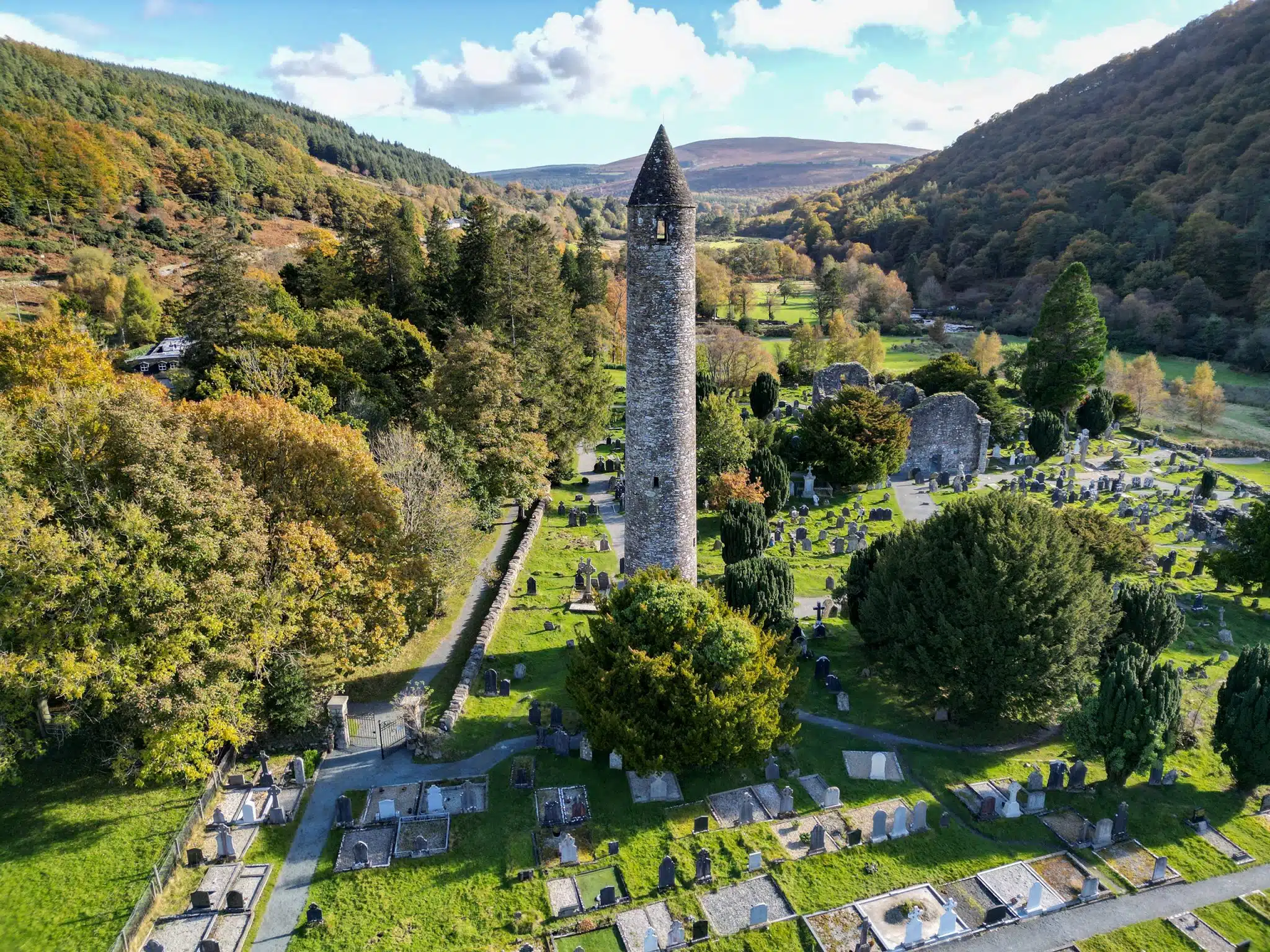 A tall, round stone tower stands in the centre of a historic cemetery with gravestones, surrounded by lush trees and rolling hills under a bright blue sky with scattered clouds.