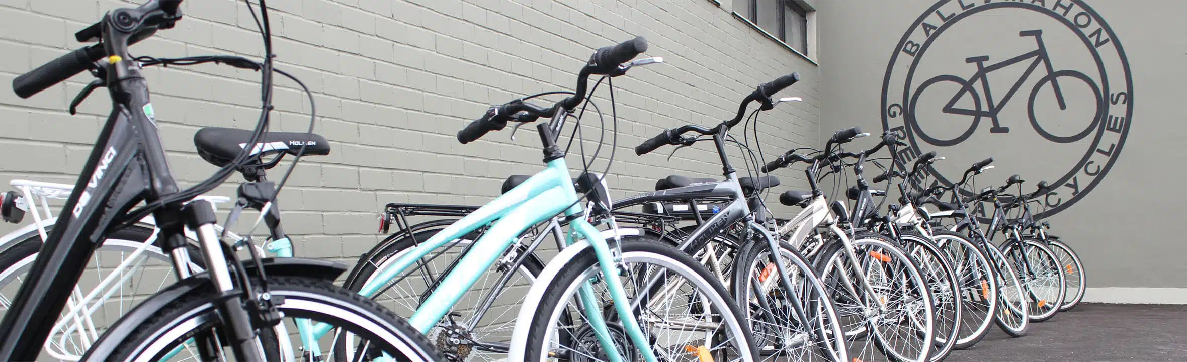 A row of bicycles is parked along a grey brick wall outside a building with a large circular bike shop logo painted on the wall.