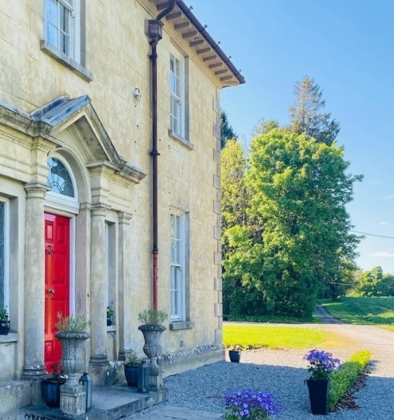 A large, light stone house with a bright red front door and classical columns. Potted plants line the entrance, and a gravel drive curves beside a green lawn and tall trees under a clear blue sky.