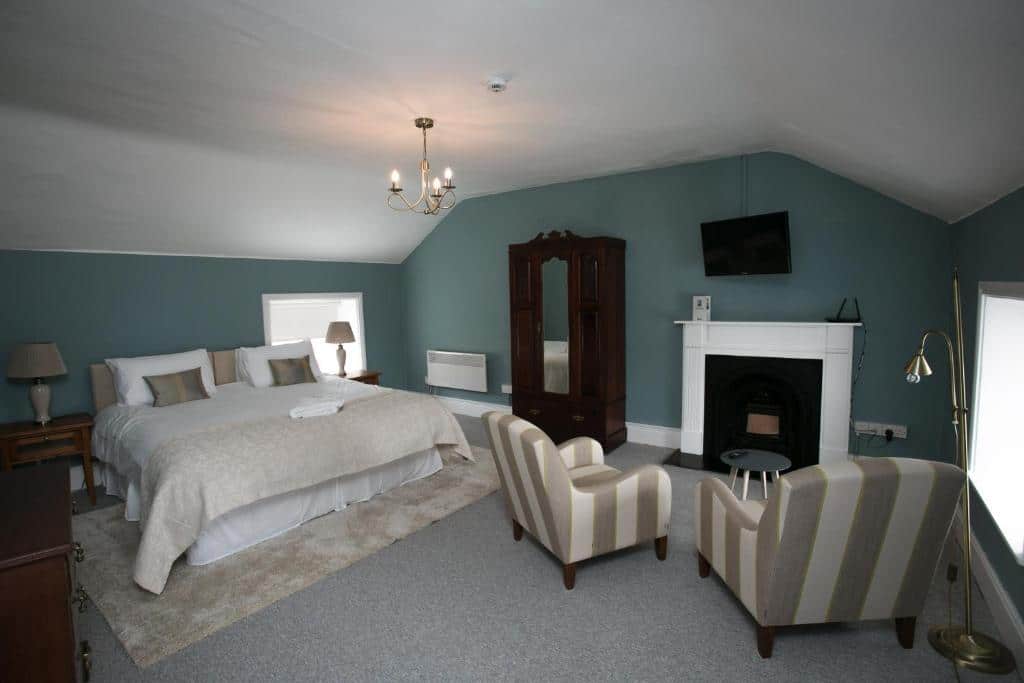 A spacious bedroom with a large bed, two striped armchairs, a wardrobe, a fireplace, a wall-mounted TV, and soft green walls. Two windows let in natural light, and neutral décor adds a cosy feel.