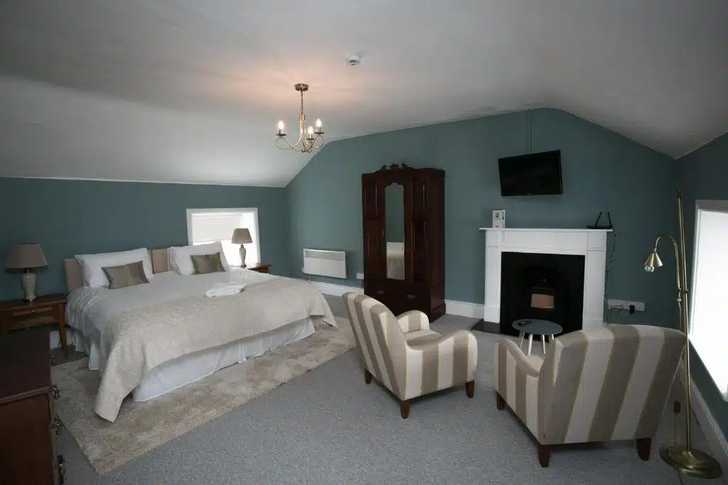 A spacious bedroom with a large bed, two striped armchairs, a wardrobe, a fireplace, a wall-mounted TV, and soft green walls. Two windows let in natural light, and neutral décor adds a cosy feel.