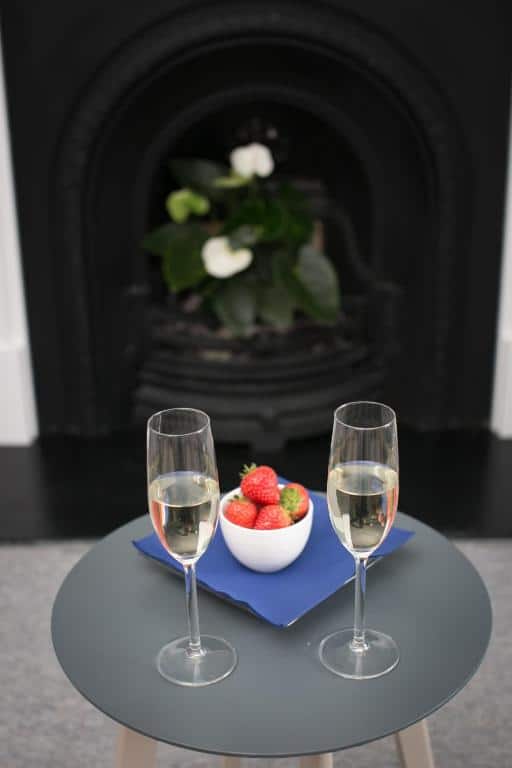 Two glasses of champagne and a white bowl of strawberries sit on a small round table with a blue serviette, in front of a black fireplace with green leafy plants and white flowers in the background.