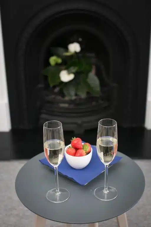 Two glasses of champagne and a white bowl of strawberries sit on a small round table with a blue serviette, in front of a black fireplace with green leafy plants and white flowers in the background.