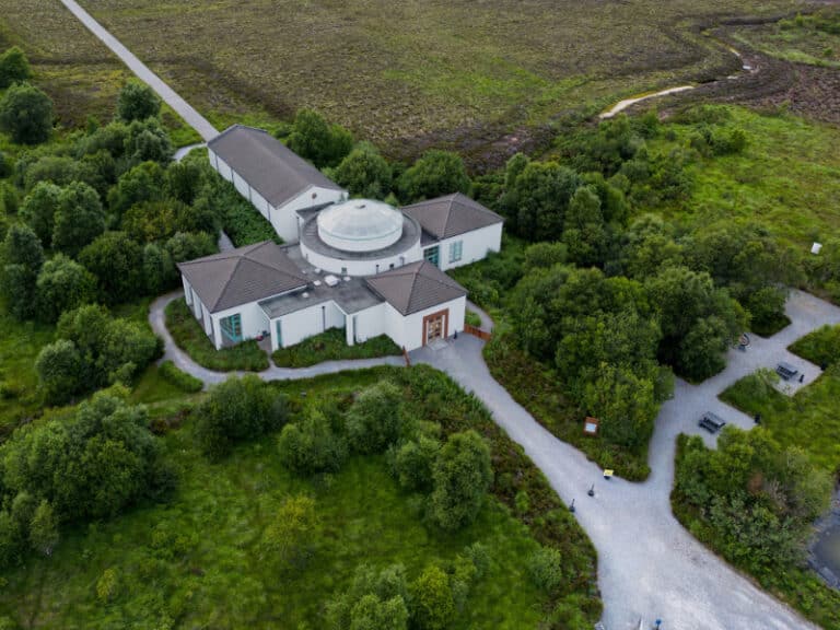 Aerial view of a white, modern building with a central glass dome surrounded by lush green trees and winding gravel paths in a rural, grassy landscape.