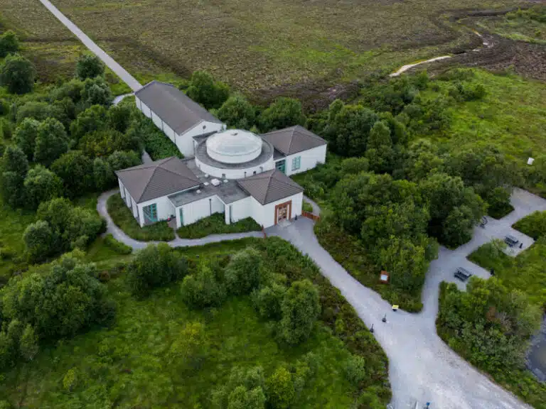 Aerial view of a white, modern building with a central glass dome surrounded by lush green trees and winding gravel paths in a rural, grassy landscape.