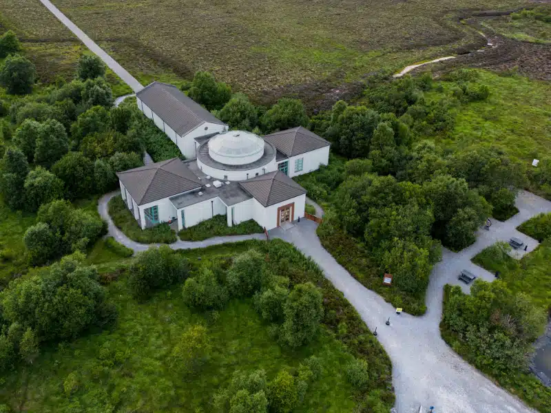 Aerial view of a white, modern building with a central glass dome surrounded by lush green trees and winding gravel paths in a rural, grassy landscape.
