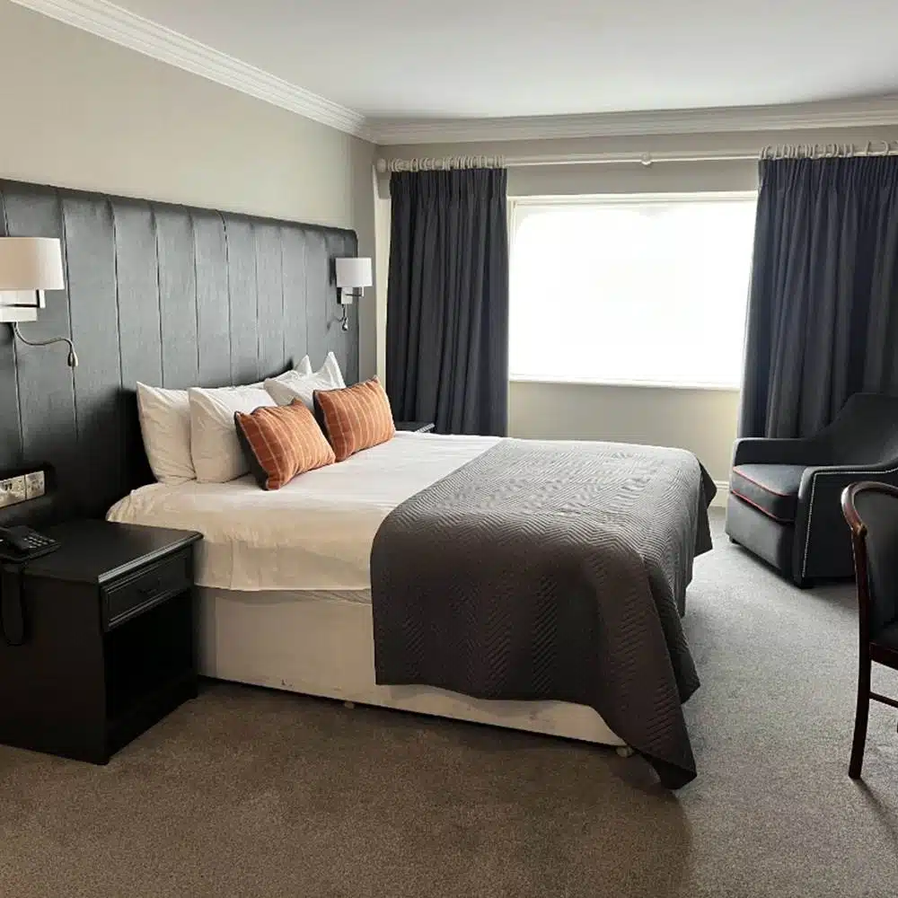 A modern hotel room with a double bed, grey headboard, orange accent pillows, dark bedside tables, a grey armchair, and a large window with dark curtains letting in natural light.