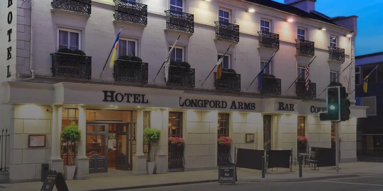 The exterior of the Longford Arms Hotel at dusk, with lit windows, several flags, decorative plants, and a sign for hotel, bar, and coffee. A traffic light and “No Parking” sign are visible in front.