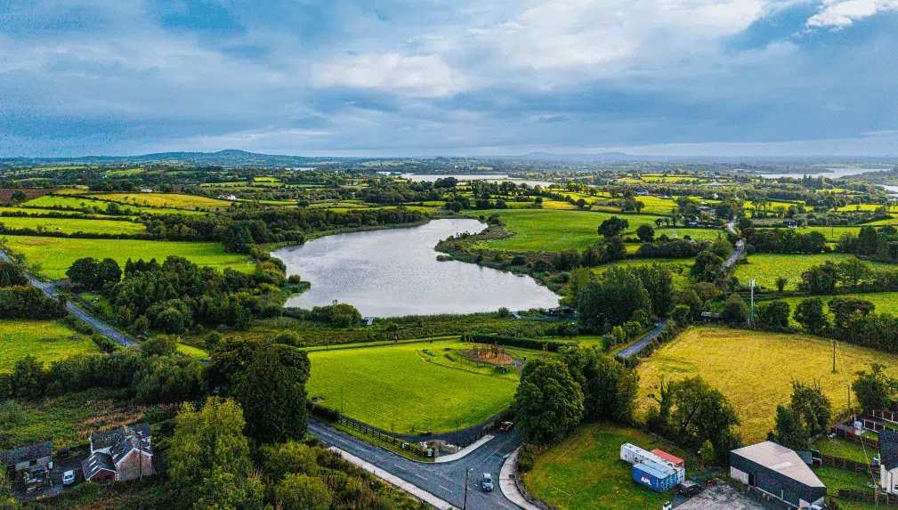 Aerial view of a lush, green countryside with patchwork fields, a large pond or lake in the centre, scattered houses, trees, and roads under a cloudy sky.