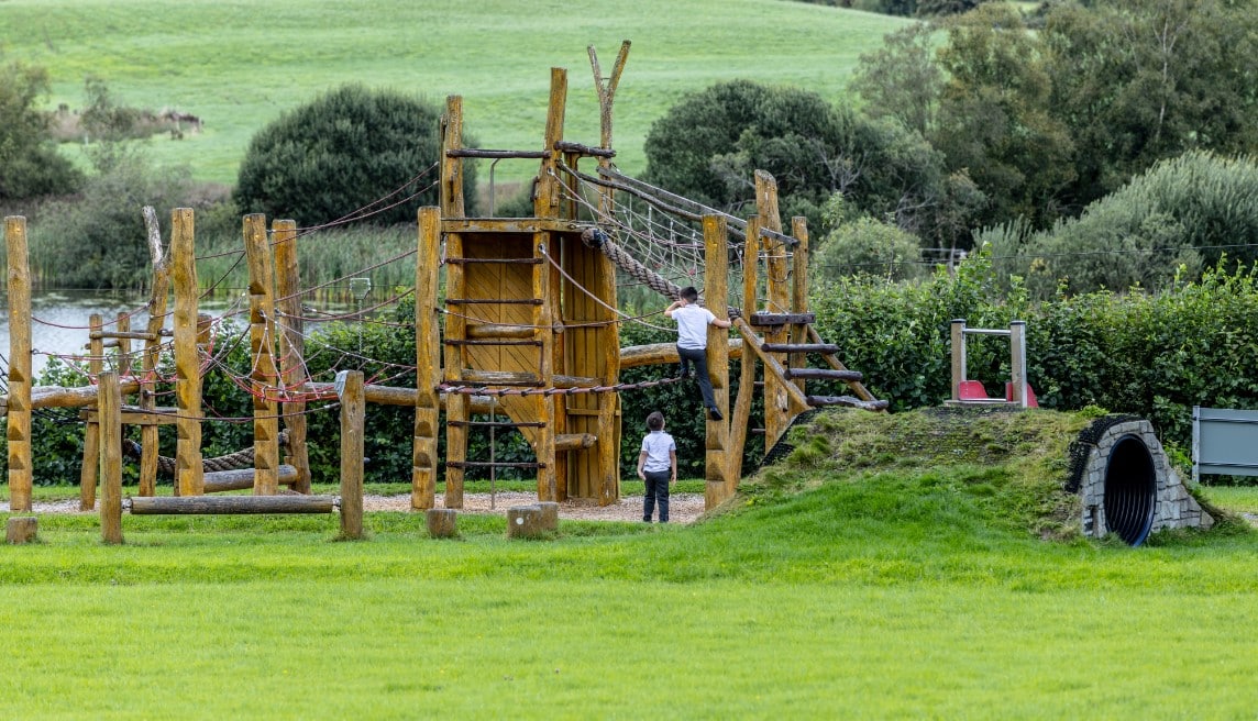 Two children play on a wooden and rope climbing structure in an outdoor playground surrounded by green grass, trees, and hills. One child climbs a wooden ladder whilst the other stands nearby watching.