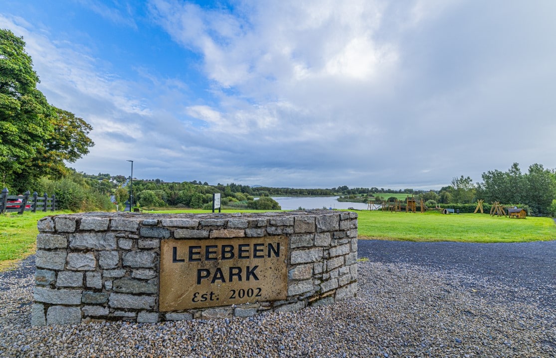 Stone entrance sign for Leebeen Park, established 2002, with a grassy park area, trees, a lake in the background, and a cloudy sky overhead.
