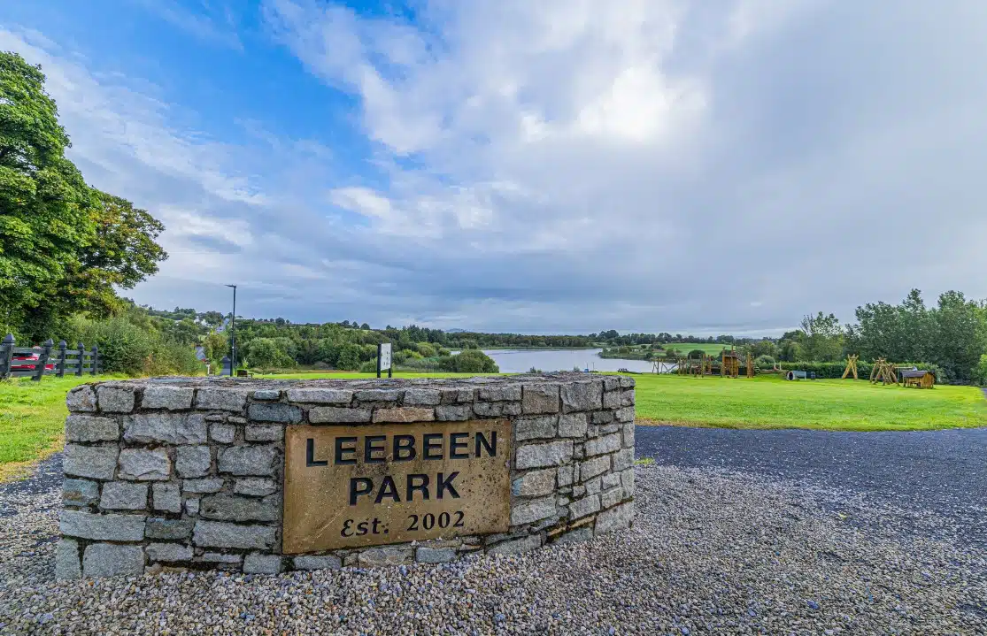 Stone entrance sign for Leebeen Park, established 2002, with a grassy park area, trees, a lake in the background, and a cloudy sky overhead.
